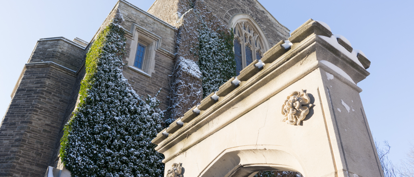 Seen from below, looking upward: The top half of Edwards Arch against the backdrop of a wall of University Hall. A dusting of snow on the arch and the ivy on the building. Blue sky.